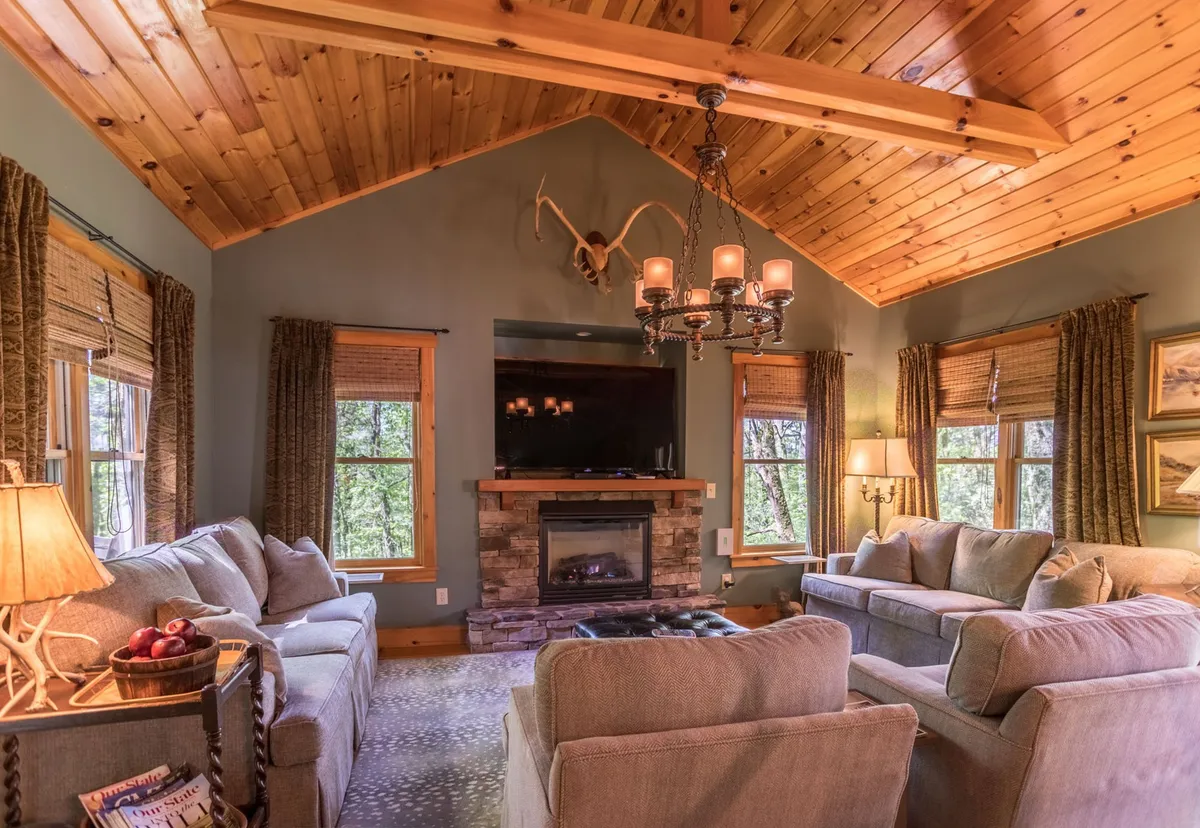 This inviting interior shot centers on a spacious living room with plush sofas arranged around a stone fireplace. A statement chandelier and antler decor contribute to the mountain cabin ambiance, and large windows fill the room with natural light. The TV is also visible, suggesting a space well-suited for movie nights or cozy gatherings.