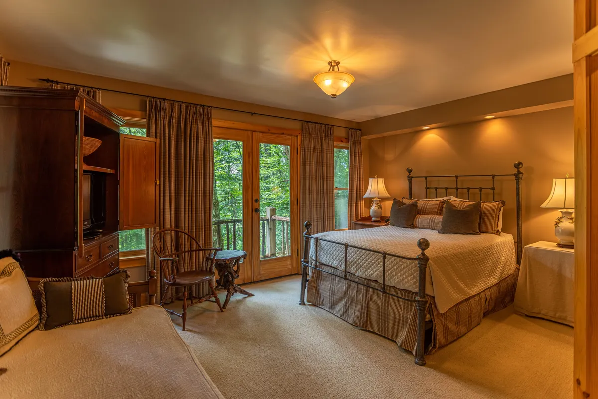 A serene bedroom with a bed adorned with plush pillows, double doors leading to a balcony, and soft, inviting textures. The setup encourages guests to enjoy the sunrise or sunset through the trees, directly connecting the indoors with the tranquil mountain landscape.