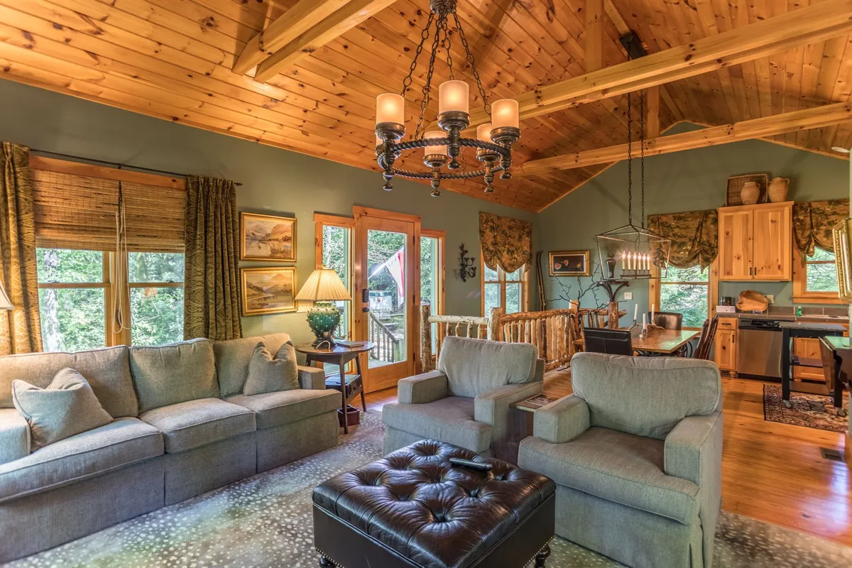 This image captures a warm, welcoming living area that seamlessly flows into the kitchen. Visible are a comfortable sofa, a striking chandelier, exposed wooden beams, the kitchen area, and large windows letting in natural light. The space is ideal for relaxing after outdoor adventures or socializing with friends and family. The open design suggests easy transitions between different activities and communal gathering spots.