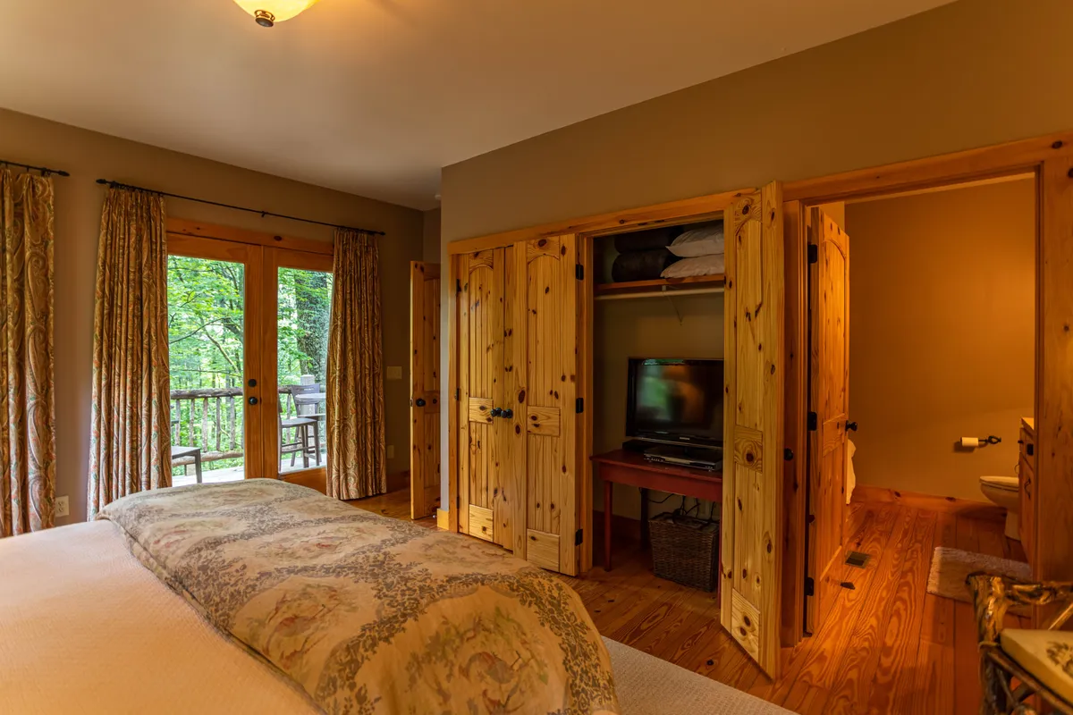 A tranquil bedroom featuring a bed with a decorative cover, natural wood accents, and double doors that open to a balcony. The doors and the view beyond hint at the natural beauty surrounding the cabin, emphasizing the connection between indoor comfort and outdoor adventure.