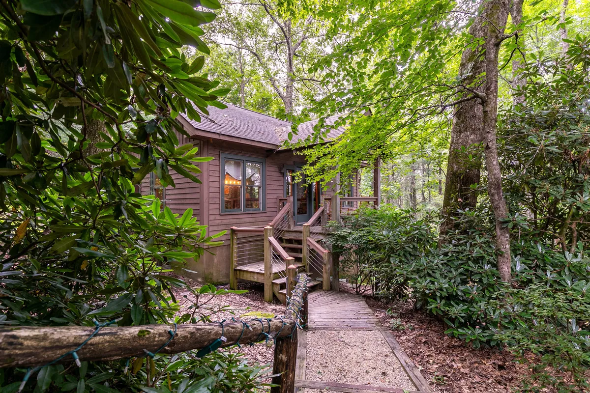 The image showcases the inviting exterior of an upscale mountain cabin, with charming wooden steps leading up to the house nestled amid lush greenery. Sunlight dances on the large windows, and a spacious porch offers a serene spot to enjoy the surrounding nature.