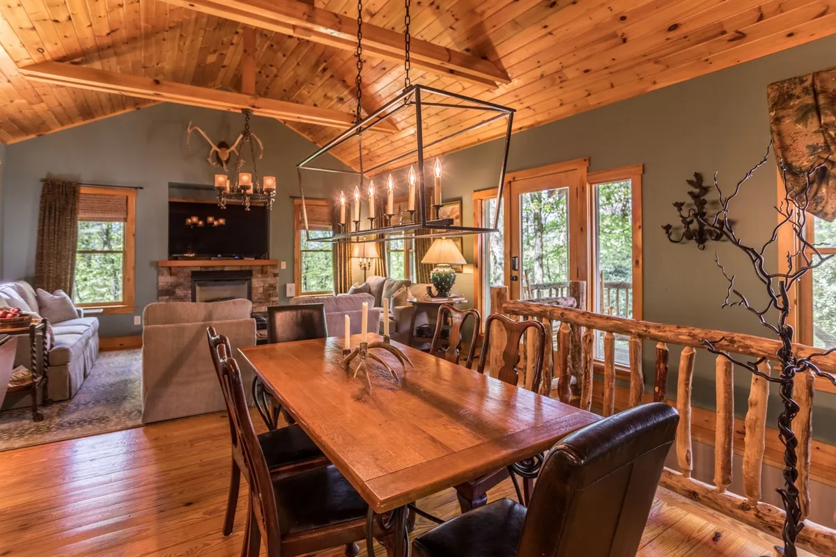 This wide-angle view captures the open-plan living and dining area, featuring a dining table set for six, a comfortable sofa, and the stone fireplace. Expansive windows offer glimpses of the surrounding forest, and the French doors leading to the deck are visible, blending indoor warmth with outdoor scenery.
