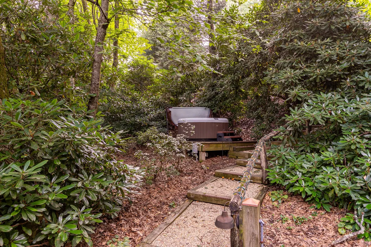 A serene wooden pathway leads to a secluded hot tub nestled among greenery. The hot tub sits on a private deck, offering an inviting spot for relaxation with views of the surrounding forest. The area feels exclusive and tranquil, ideal for stargazing or soaking on crisp evenings.