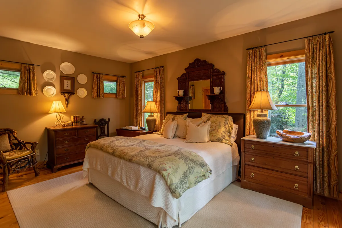 A tranquil bedroom featuring a king-sized bed with decorative bedding, wooden dressers, a window allowing in natural light, and a decorative mirror. The space feels airy, comfortable, and is clearly designed for relaxation and restful moments.