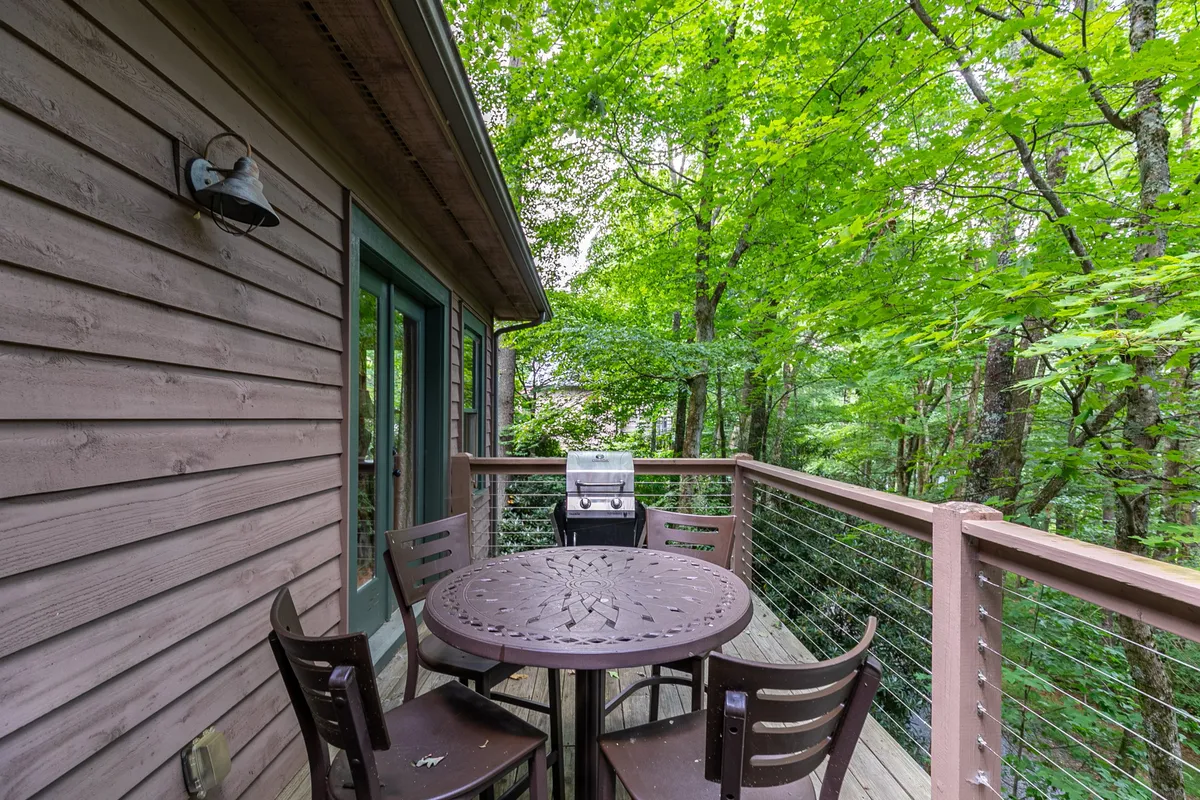 An exterior shot of a round table with four chairs and a modern grill, all set on a deck surrounded by lush green trees. The setup encourages outdoor dining, barbecuing, and socializing in a serene, natural environment. The grill is prominently visible, underscoring the space's suitability for evening barbecues.