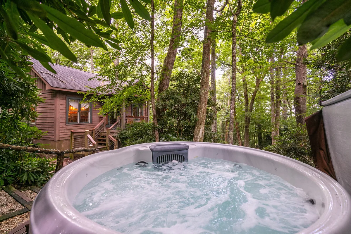 The exterior of a mountain cabin featuring a hot tub on a wooden deck, surrounded by towering trees. The cabin itself is visible, set against the natural landscape, conveying a sense of privacy and relaxation.