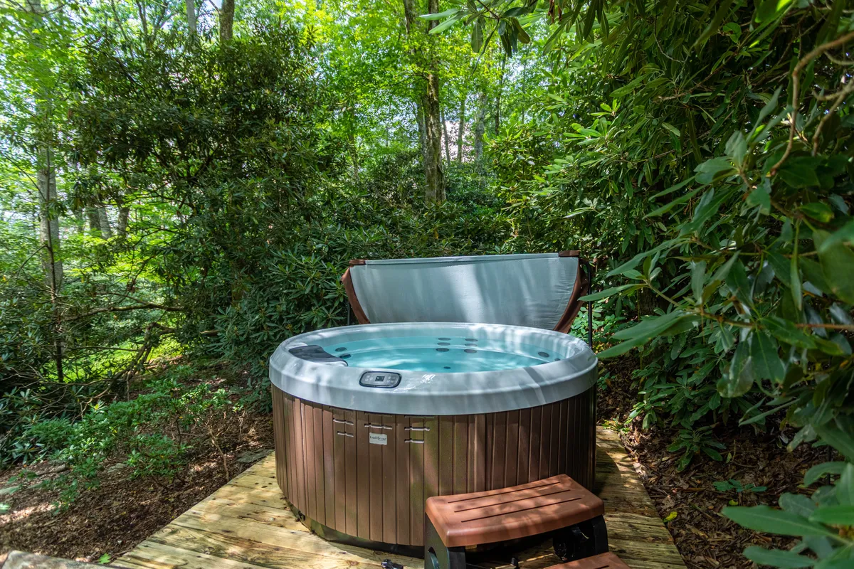 This exterior image features a spacious wooden deck with a hot tub, surrounded by lush greenery. The deck includes a comfortable bench, inviting guests to unwind while being immersed in nature. The view highlights the tranquility and outdoor relaxation opportunities available at the cabin.